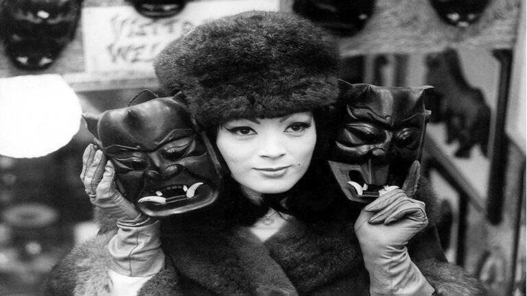 Image of Tura Satana in a fur hat holding up two Japanese hannya masks.