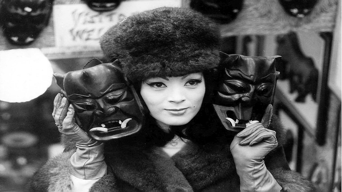 Image of Tura Satana in a fur hat holding up two Japanese hannya masks.